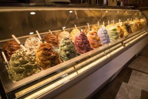 History of gelato: Photo of artificial commercial gelato case filled inauthentic gelato created with lab-created ingredients-gelato is in piles high above the rim of their tubs, with toppings and syrups.